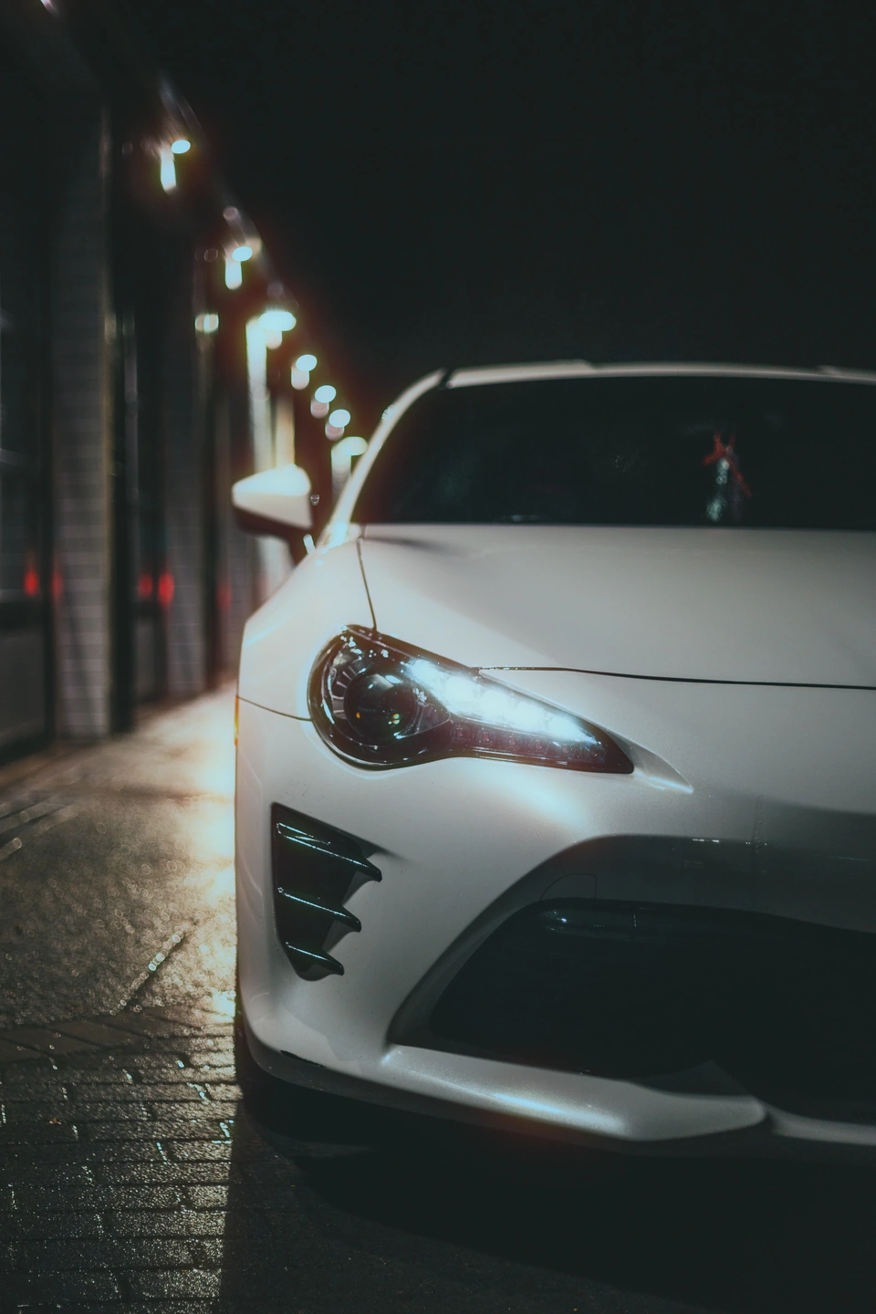 White sports car at night, with warm halos around the headlight and a row of street lights