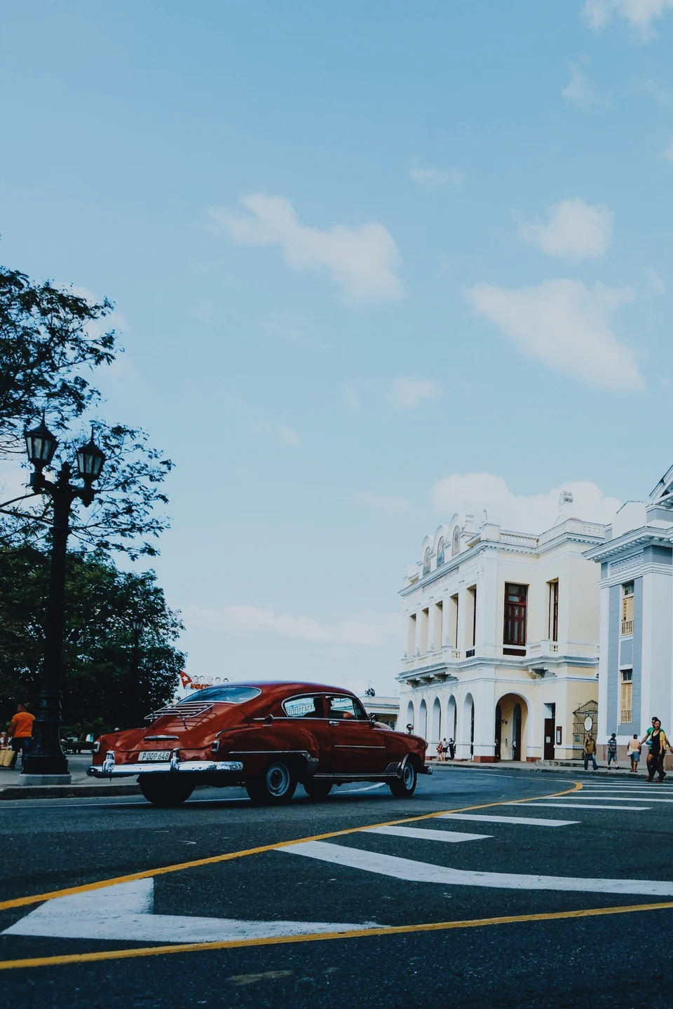 A red vintage car passing pale city buildings beneath a blue sky, with the City Chrome preset in Photonica